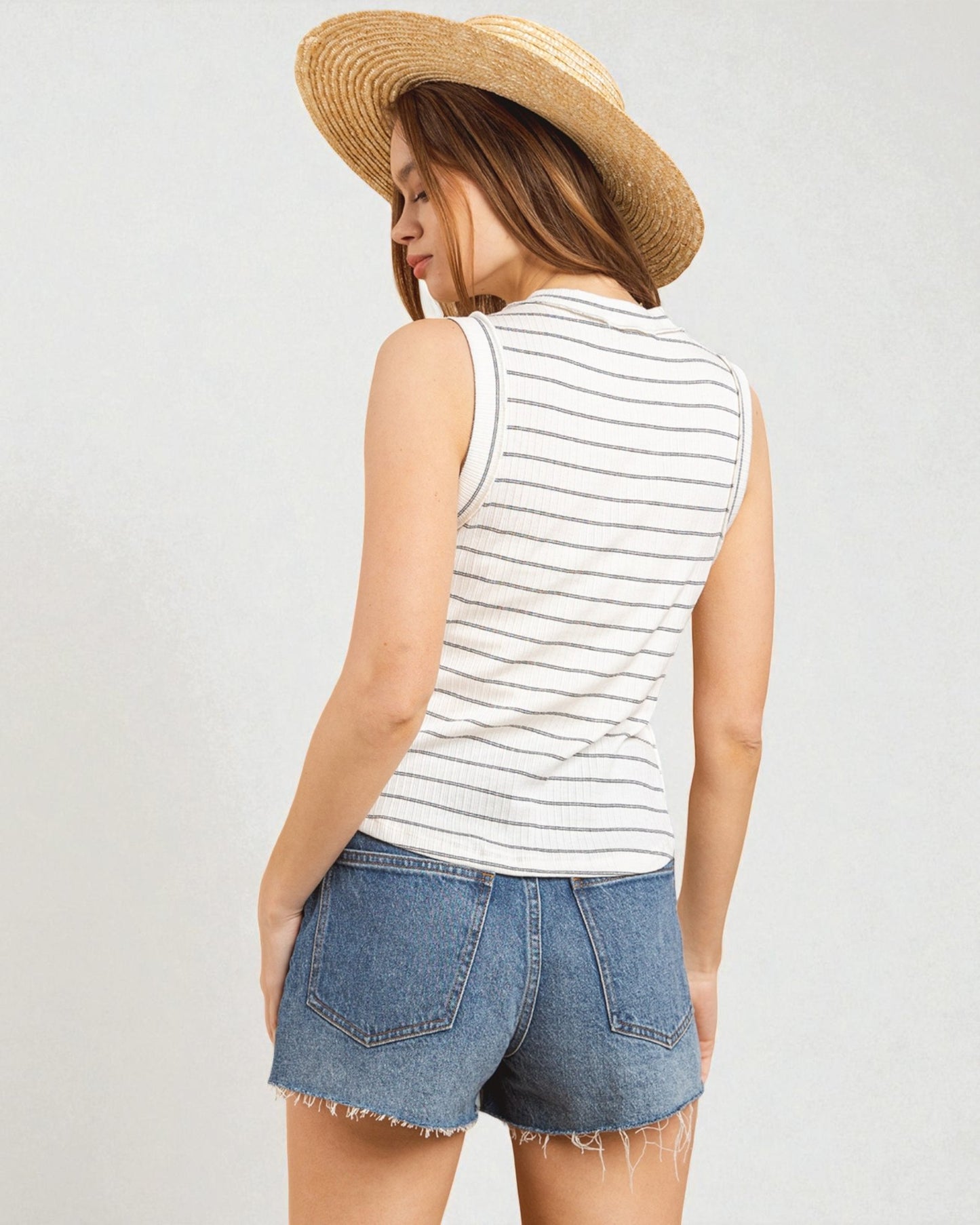Woman wearing a striped tank top and denim shorts with a straw hat on a light gray background