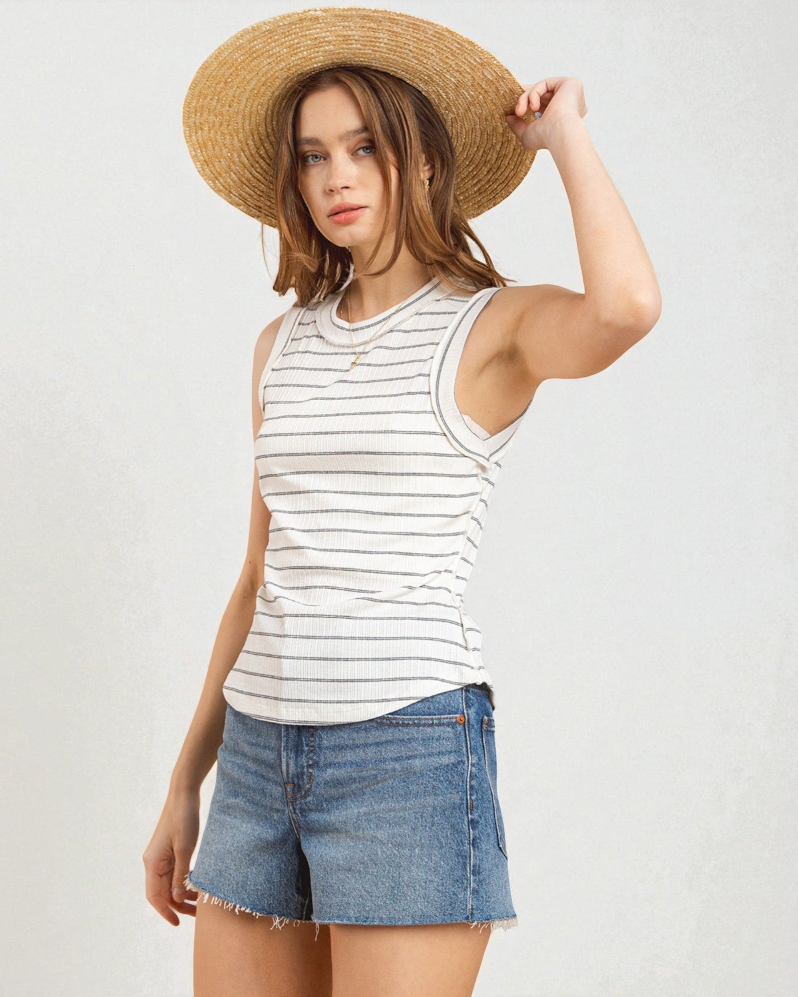 Woman wearing a striped tank top, denim shorts, and a straw hat on a plain background