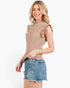 A woman modeling a sleeveless beige knit top with ruffle trim on the cap and shoulders, paired with distressed denim shorts, against a plain white background.