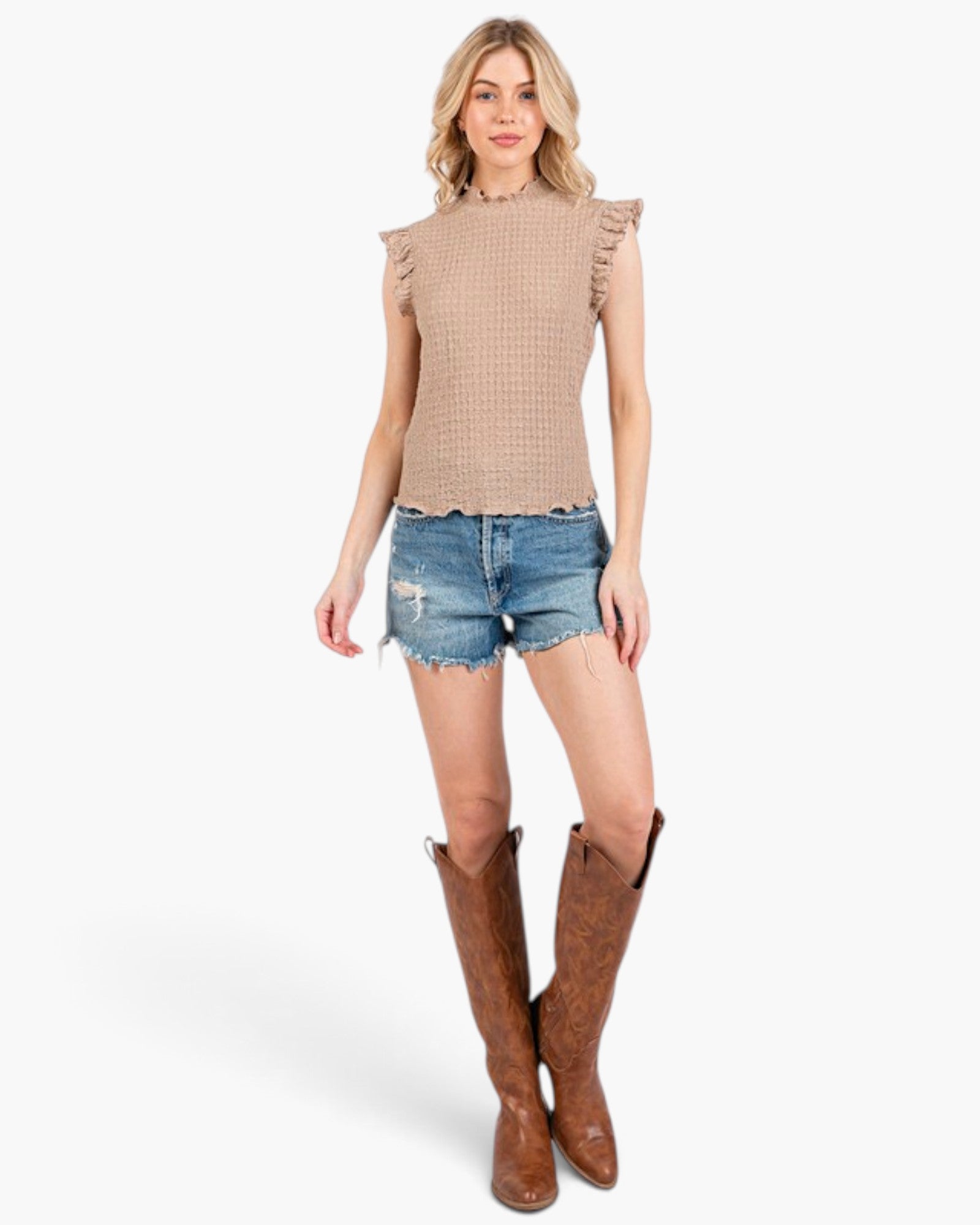 A woman modeling a sleeveless beige knit top with ruffle details on the shoulders, paired with distressed denim shorts and brown cowboy boots, against a plain white background.