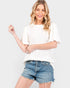 A woman modeling a white short-sleeve textured top with ruffled sleeves and blue denim shorts against a light gray studio background.