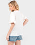 A woman wearing a white textured short-sleeve blouse with ruffled sleeves and denim shorts, shown from a three-quarters back view against a light gray studio background.