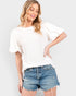 A woman wearing a white textured short-sleeve top with flutter sleeves and distressed blue denim shorts, posing against a light gray studio background.