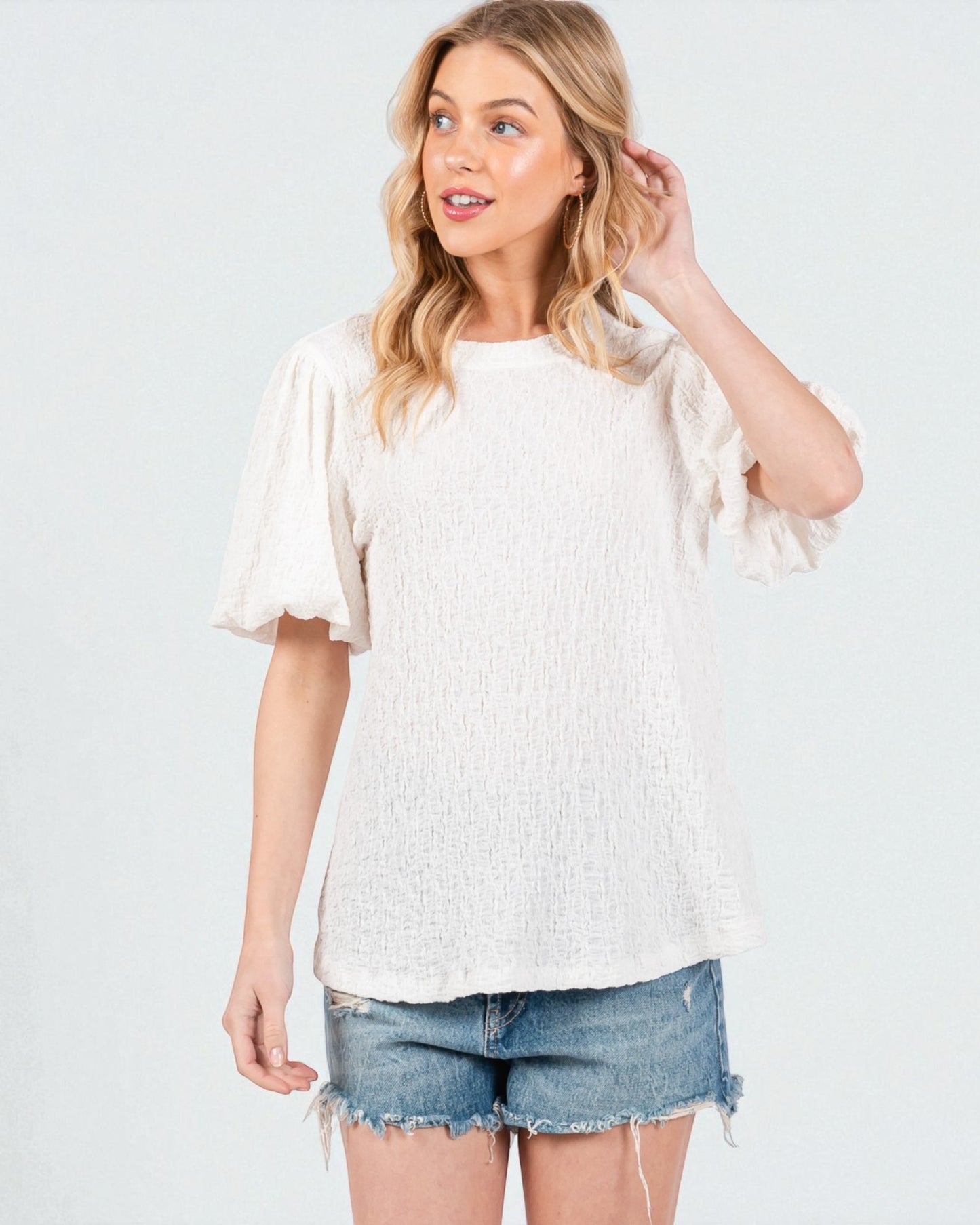 Woman wearing a white textured blouse and denim shorts on a light gray background