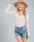Woman wearing a white lace long-sleeve top and denim shorts with a beige hat on a gray background