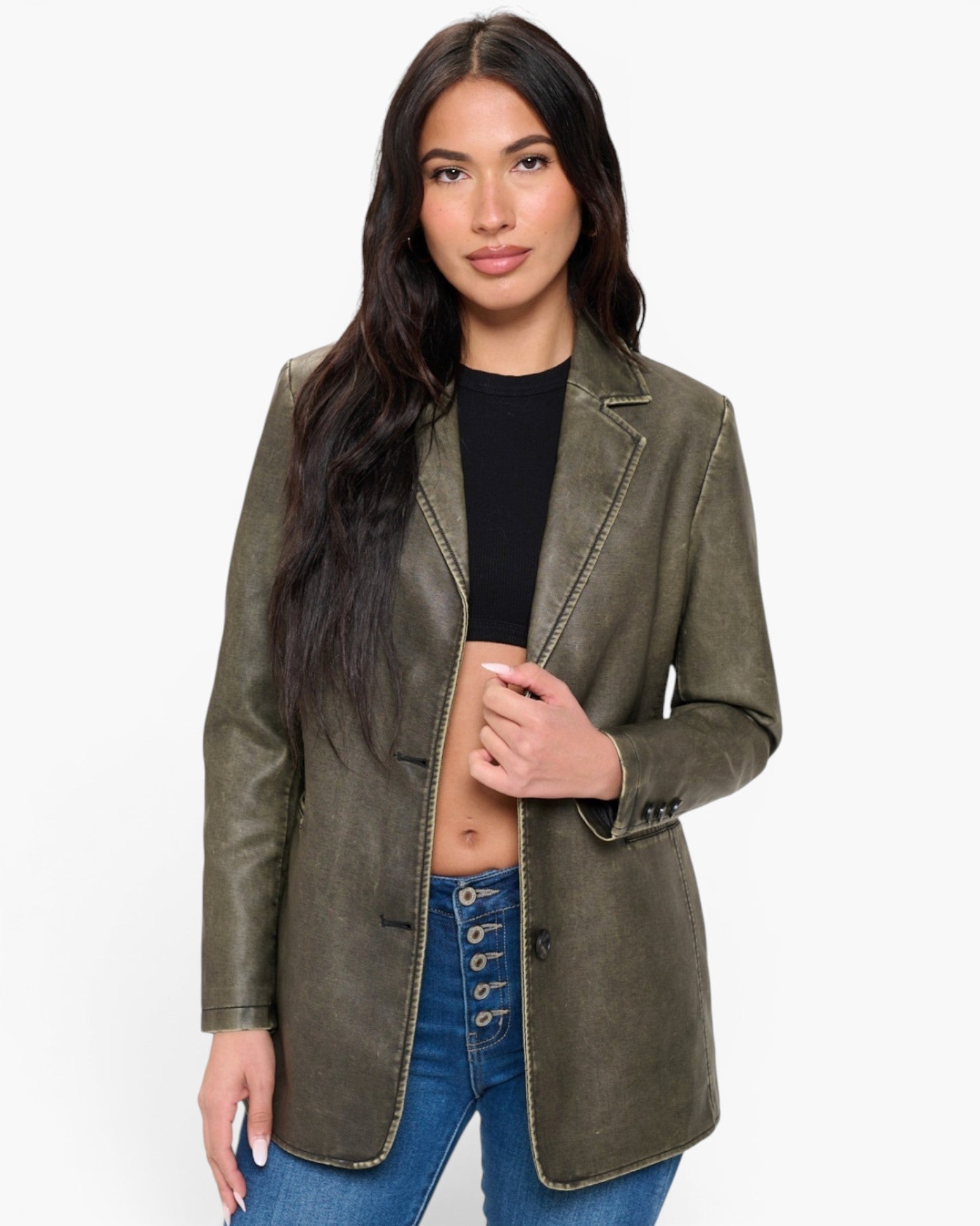 A woman modeling an olive green faux-leather blazer with contrast gold-toned stitching, worn over a black crop top and blue jeans against a plain white background.