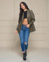 Woman wearing a green jacket, black top, and blue jeans against a beige wall.