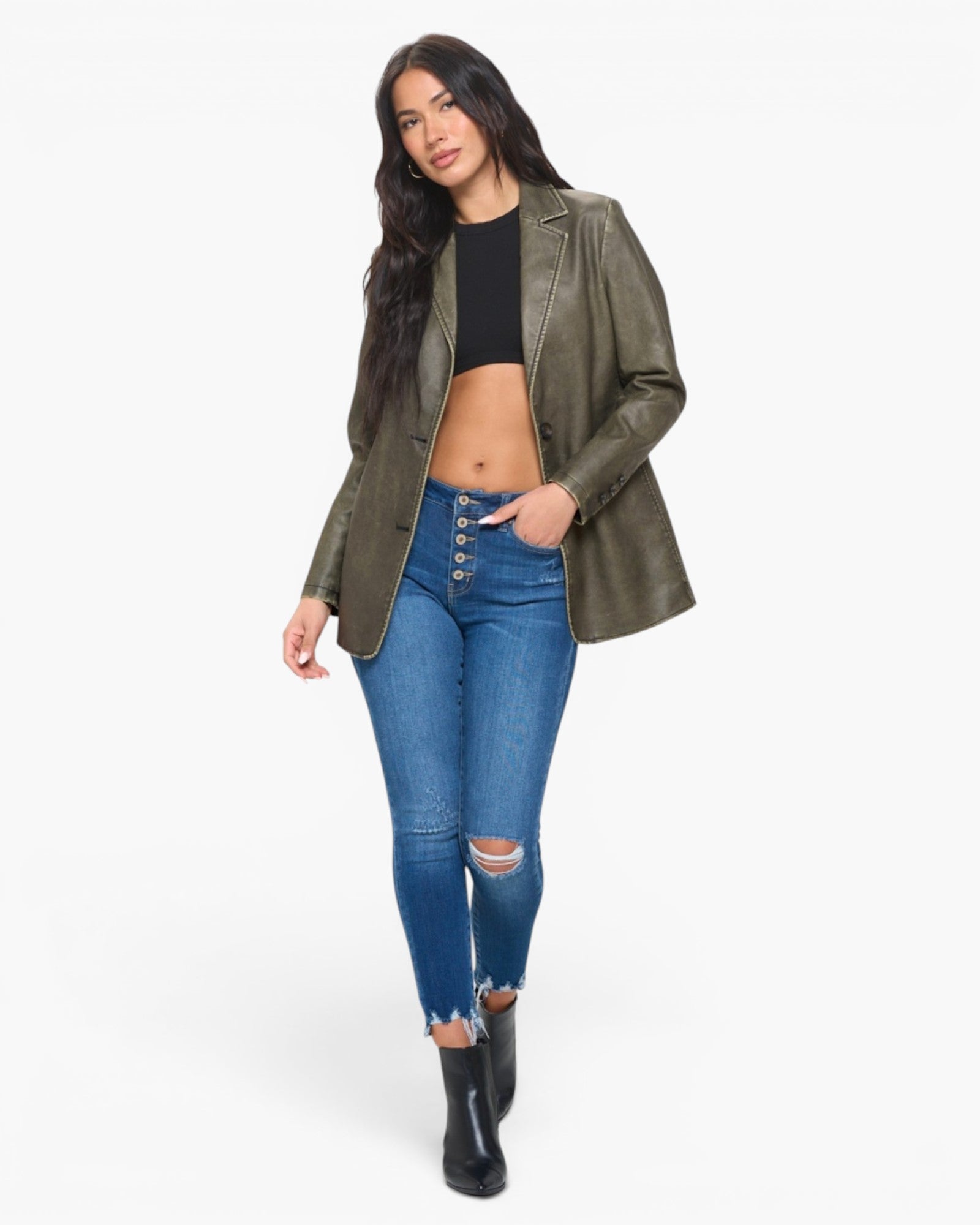 A woman modeling an olive green faux-leather blazer over a black crop top and blue distressed jeans, standing against a plain white studio background.