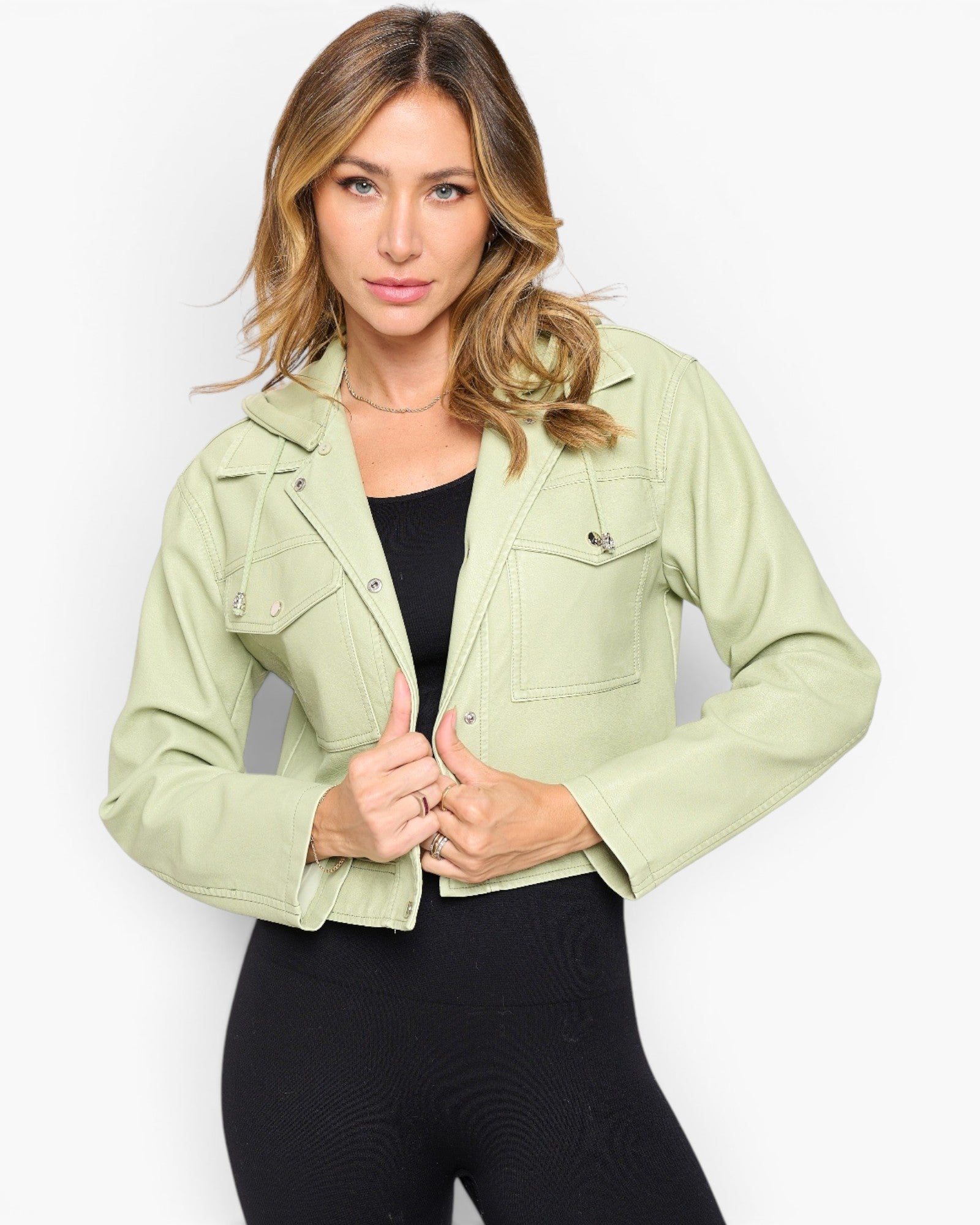 A woman modeling a light sage green cropped denim jacket over a black top and black leggings, set against a plain light gray studio background.