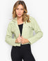 A woman modeling a light sage green cropped denim jacket over a black top and black leggings, set against a plain light gray studio background.
