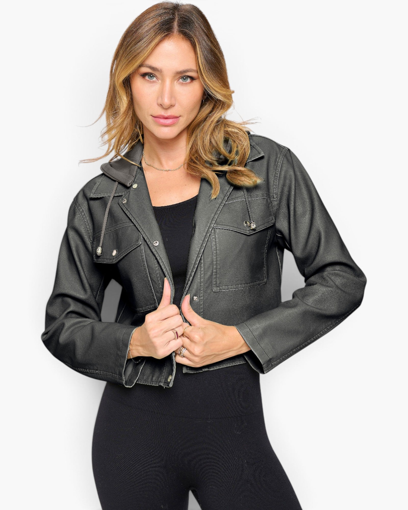 A woman modeling a cropped black faux-leather jacket with silver snap details, worn over a black top and black leggings, against a light gray studio background.