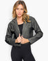A woman modeling a cropped black faux-leather jacket with silver snap details, worn over a black top and black leggings, against a light gray studio background.