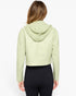 A woman viewed from the back wearing a light sage green cropped hooded jacket and black leggings against a plain white studio background.