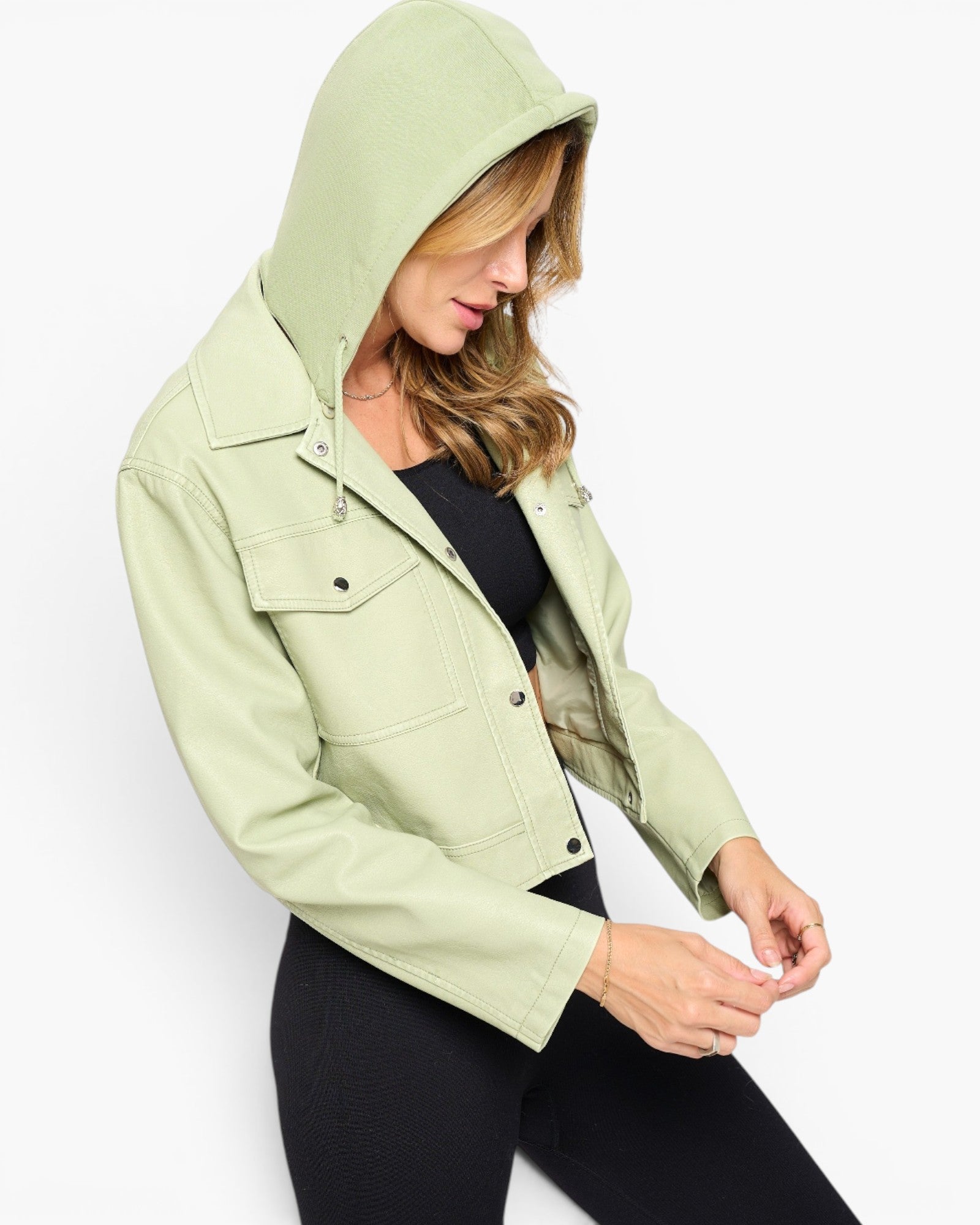 A woman wearing a light sage green hooded jacket over a black top and black pants, photographed against a clean white studio background.