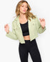 A woman modeling a cropped sage green hooded jacket over a black sports bra and black leggings, photographed against a clean white studio background.