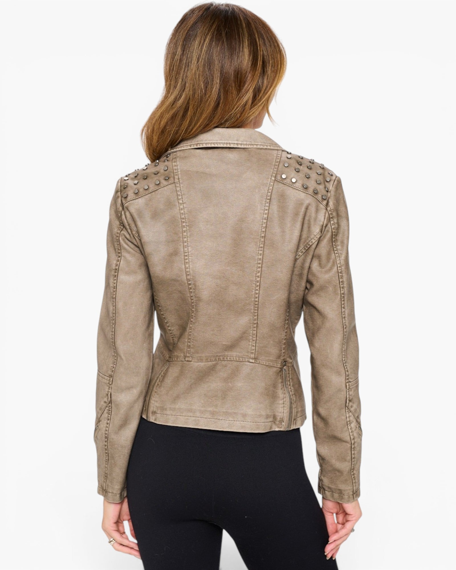 A woman viewed from the back wearing a cropped beige faux-leather jacket with silver studs on the shoulders, paired with black pants, against a plain white background.