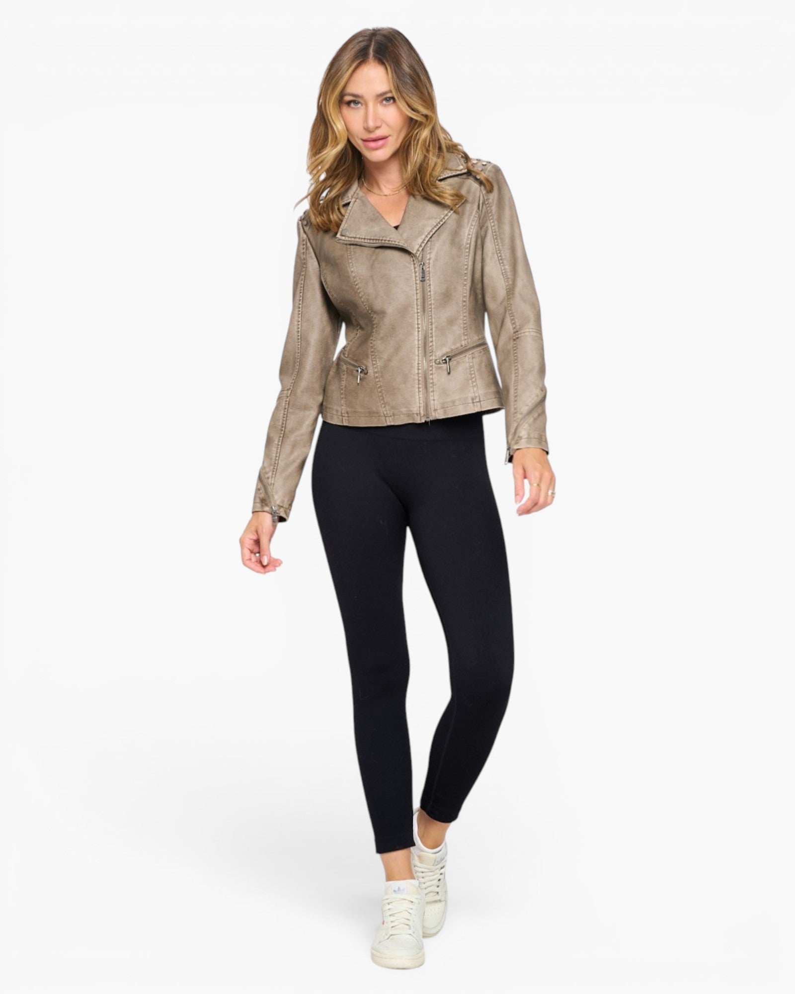 A woman modeling a cropped, asymmetrical zip-front moto jacket in a textured taupe-brown faux leather, paired with black leggings and white sneakers, against a solid white background.