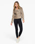 A woman modeling a cropped, asymmetrical zip-front moto jacket in a textured taupe-brown faux leather, paired with black leggings and white sneakers, against a solid white background.
