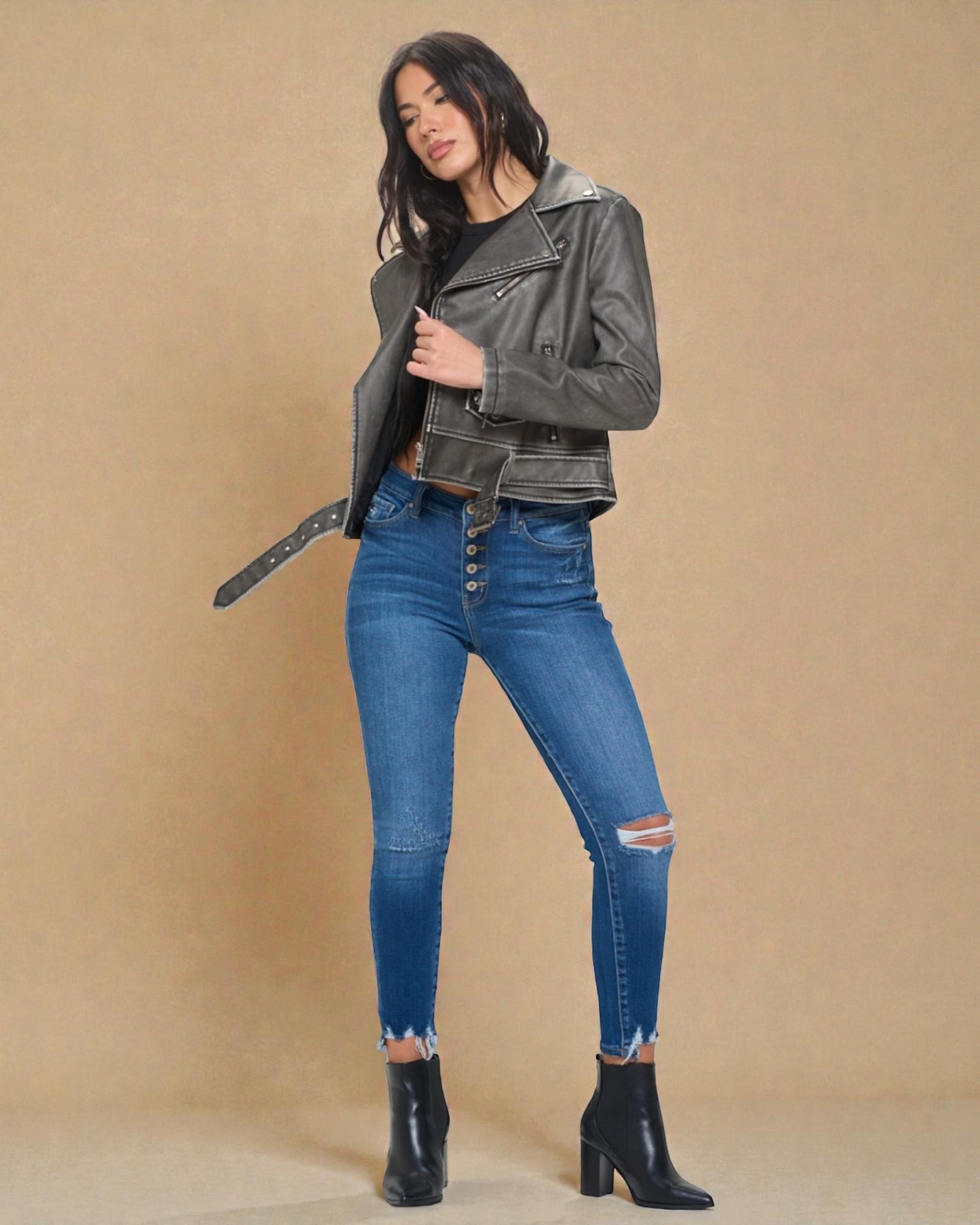 Woman wearing a gray leather jacket, blue jeans, and black boots on a beige background
