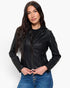 A woman modeling a fitted black faux-leather moto jacket over a black top and blue jeans, photographed against a plain light gray studio background.