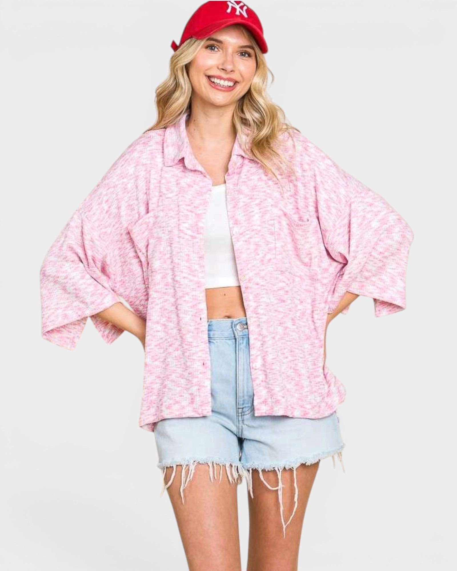 Woman wearing a pink patterned shirt, white crop top, and denim shorts with a red cap on a light gray background