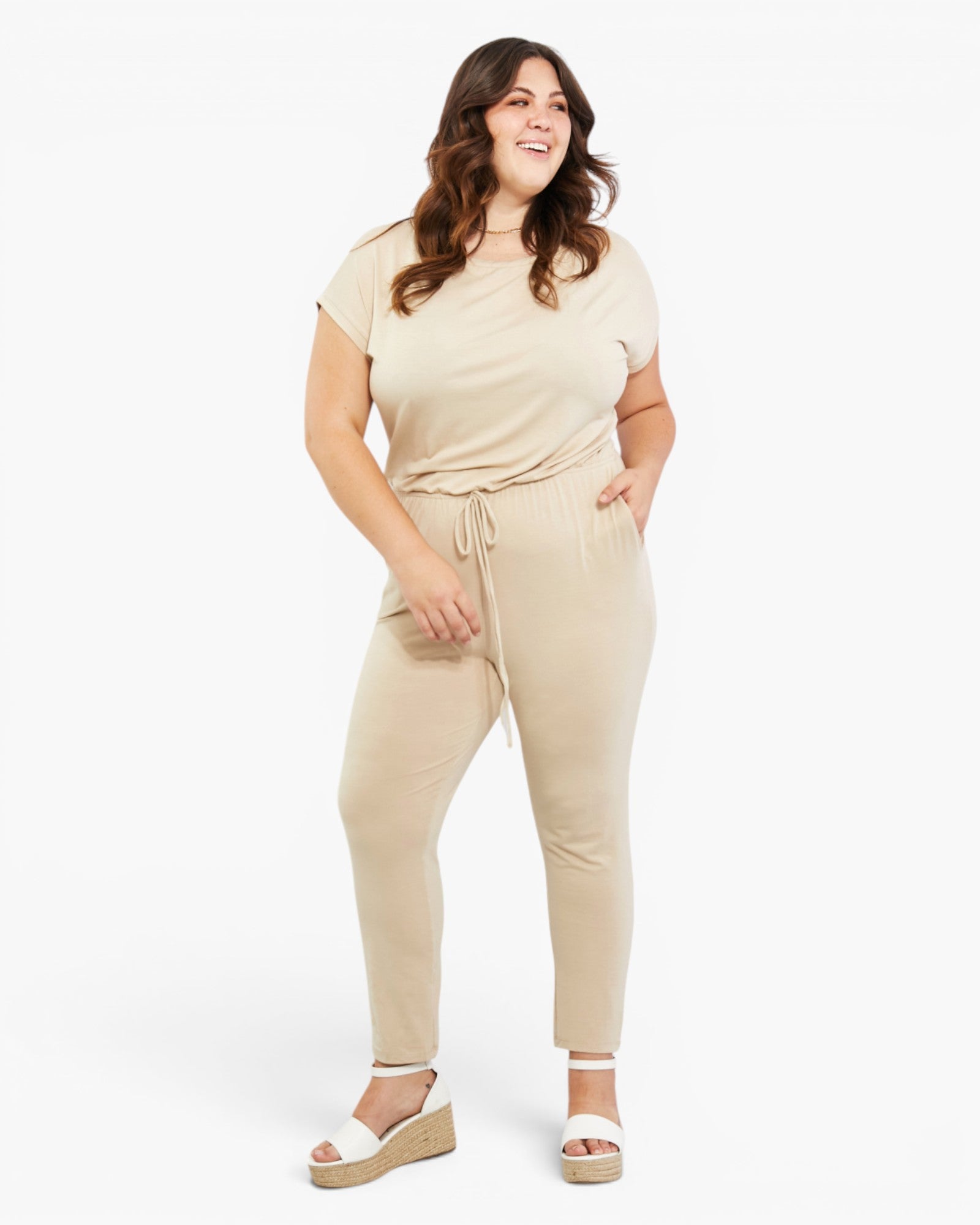 A woman modeling a beige short-sleeve t-shirt and matching drawstring pants set with white wedge sandals, photographed against a clean white studio background.