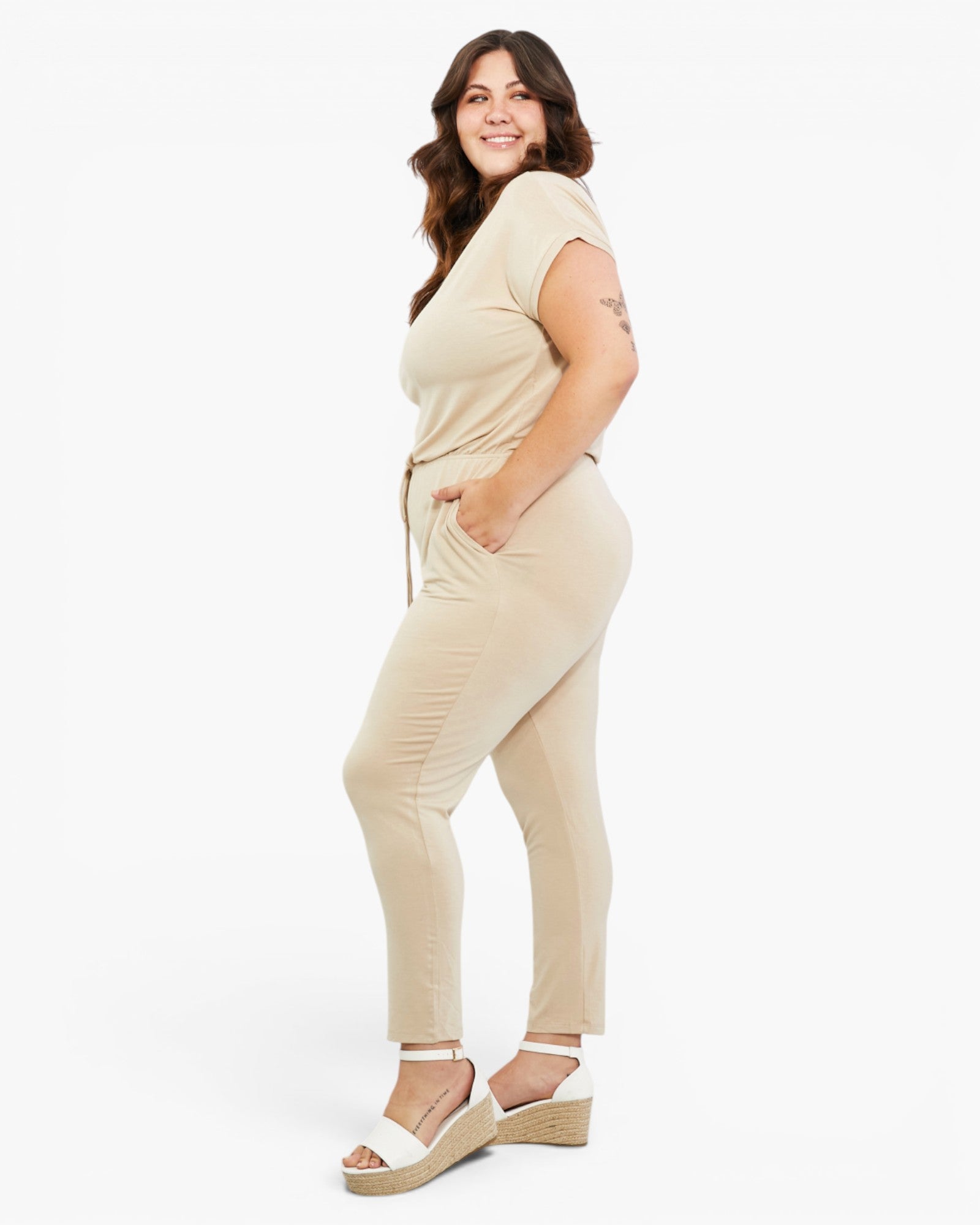 A woman modeling a beige short-sleeve jumpsuit with a matching tie belt, paired with white wedge sandals, against a plain white studio background.