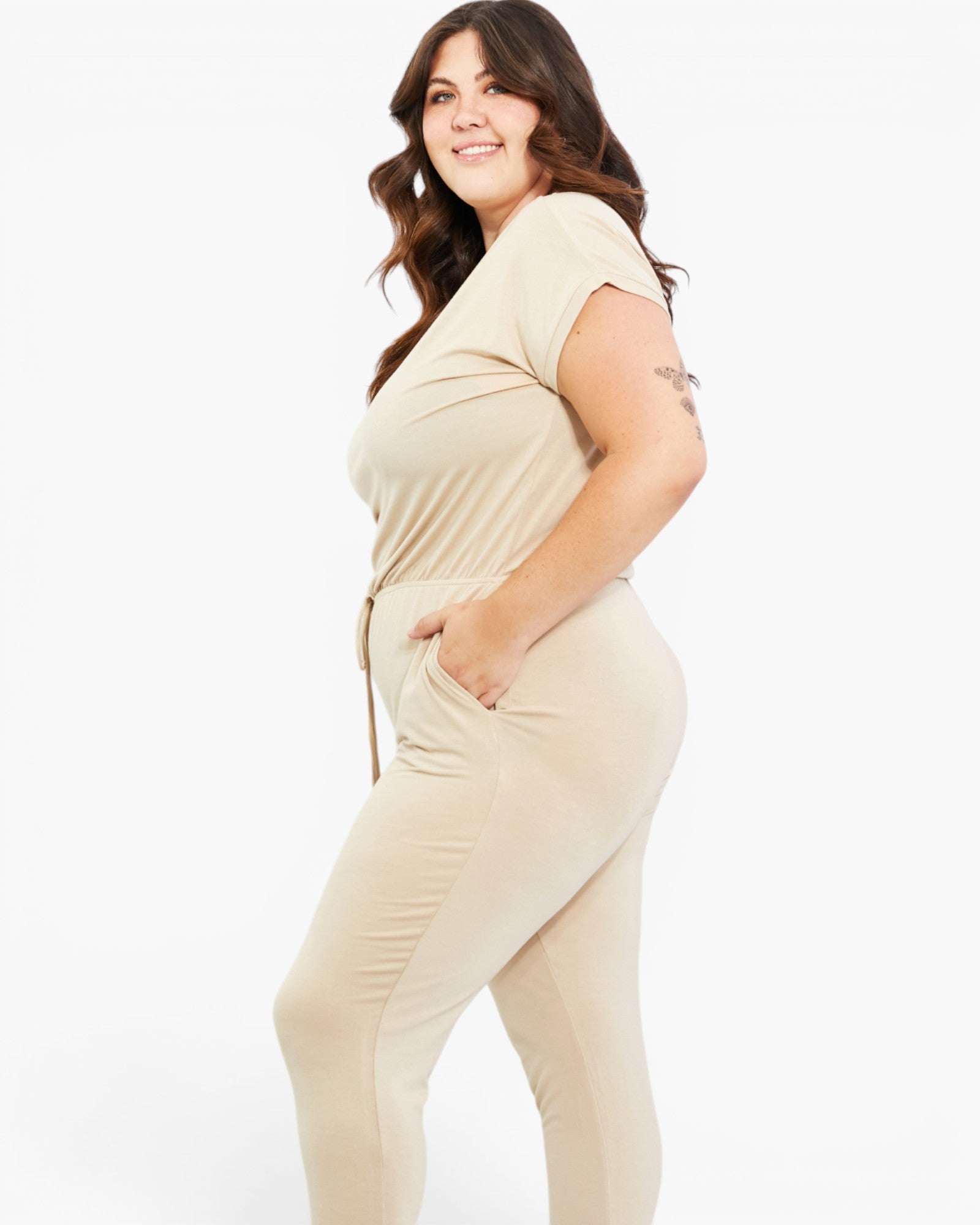 A woman modeling a beige short-sleeve jumpsuit with side pockets and a drawstring waist, photographed against a plain white studio background.