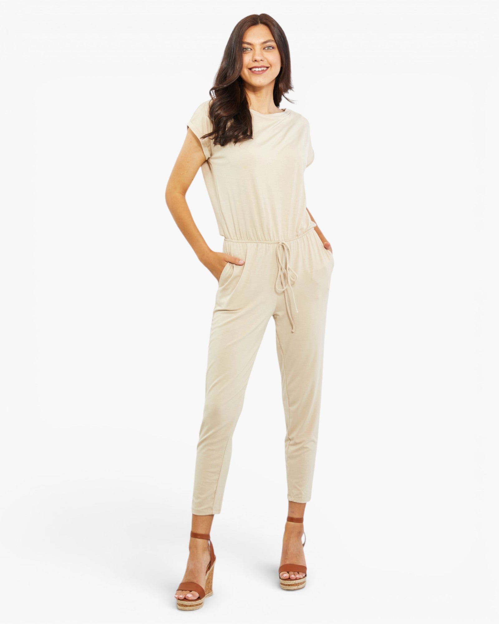 A woman modeling a light beige short-sleeve jumpsuit with a drawstring waist and side pockets, paired with brown wedge sandals, against a plain white studio background.