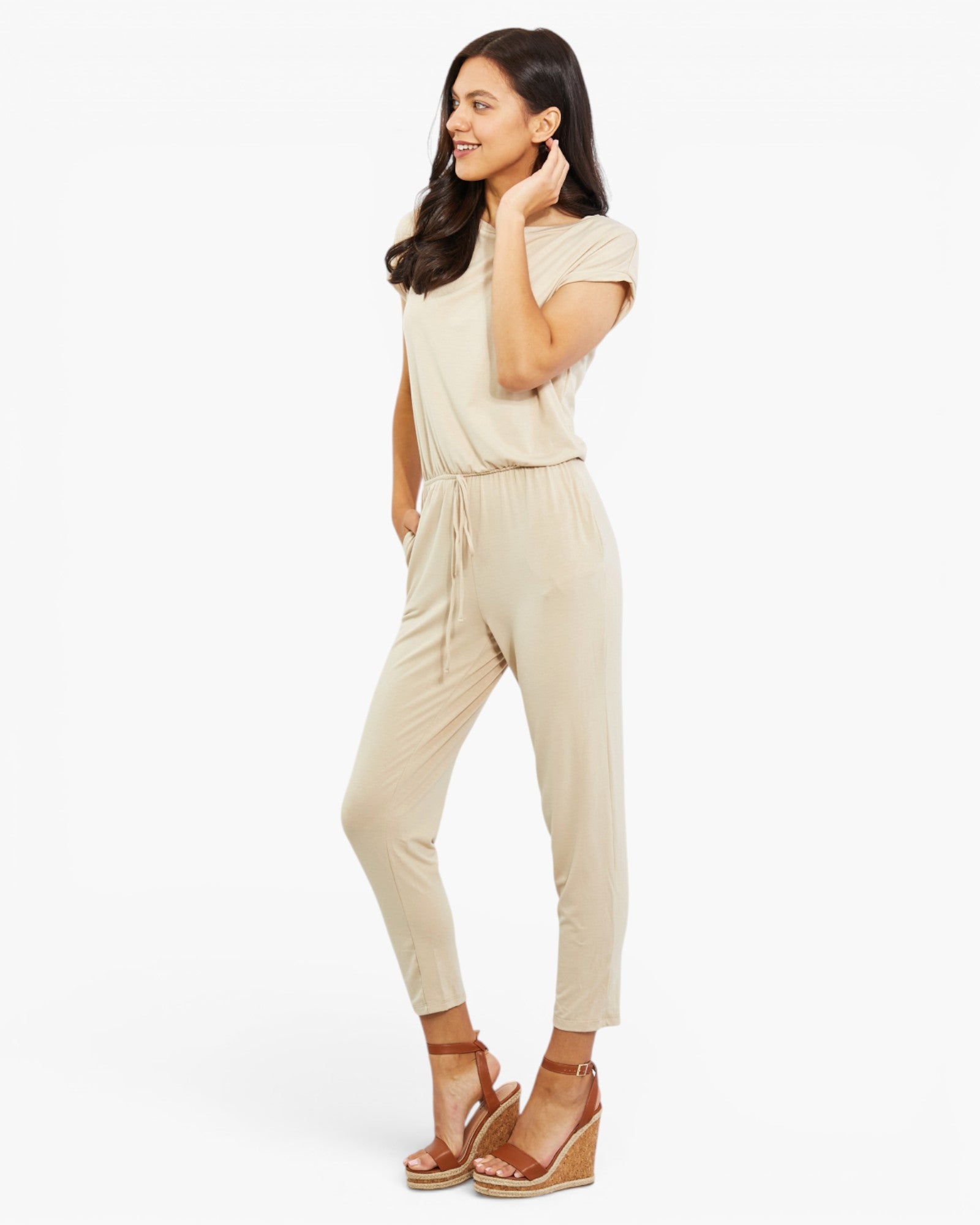 A woman modeling a light beige short-sleeve jumpsuit with a drawstring waist, paired with brown wedge sandals, against a plain white studio background.