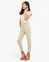 A woman modeling a light beige short-sleeve jumpsuit with a drawstring waist, paired with brown wedge sandals, against a plain white studio background.