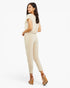 A woman modeling a light beige short-sleeve jumpsuit with a relaxed fit and ankle-length legs, shown from a three-quarters back angle against a plain white studio background.