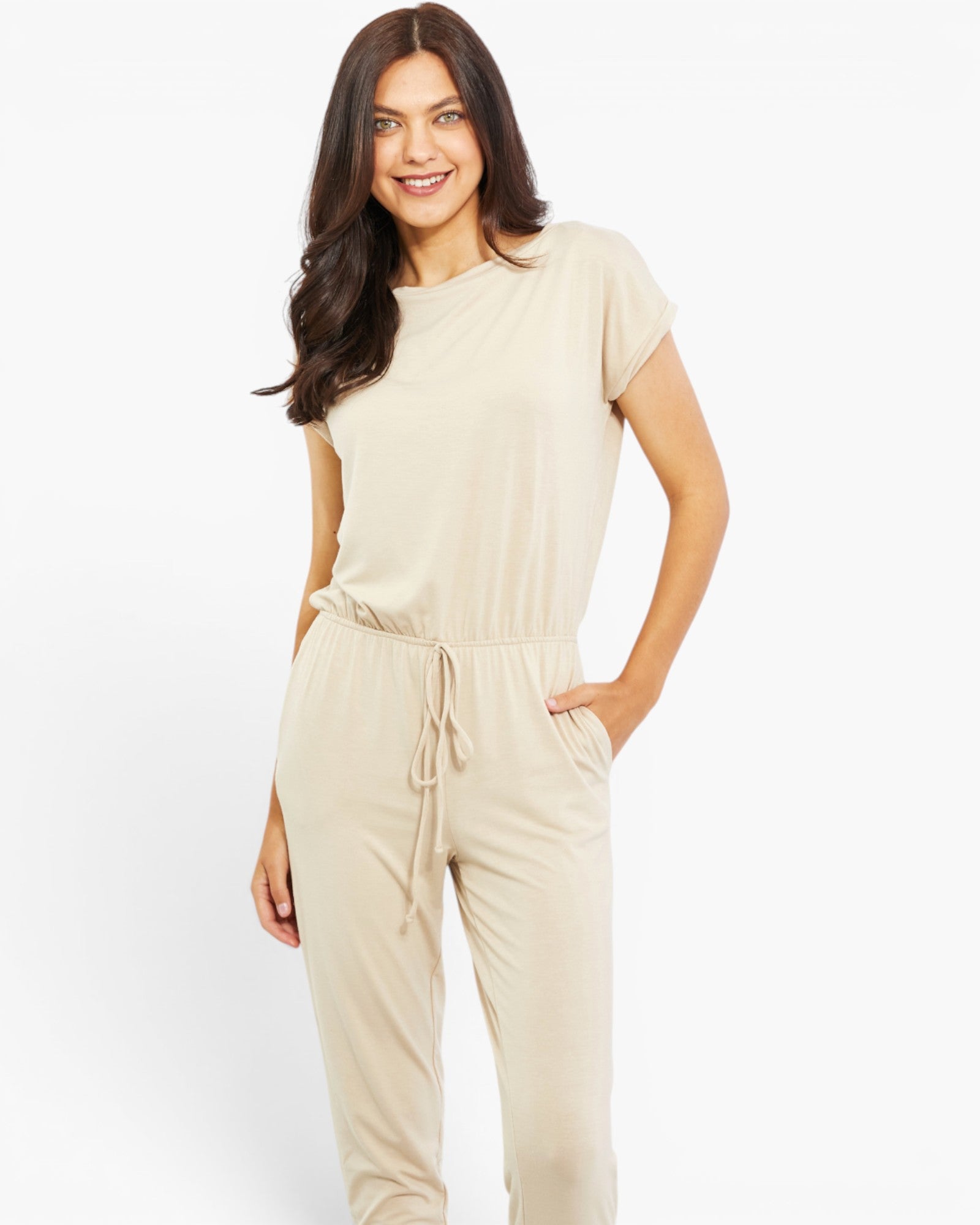 A woman modeling a light beige short-sleeve jumpsuit with a drawstring waist and side pockets, photographed against a clean white studio background.