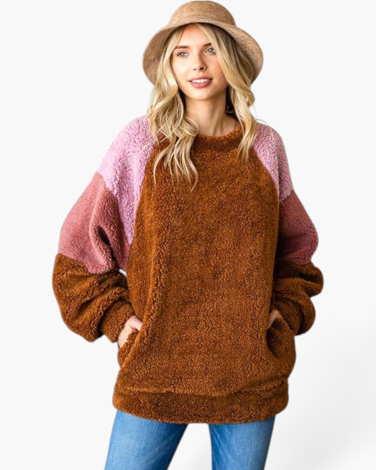 A woman wearing a fuzzy colorblock pullover sweater with a brown body, pink sleeves, and a brown bucket hat, paired with blue jeans, against a plain white background.