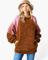 A woman wearing a fuzzy colorblock pullover sweater with a brown body, pink sleeves, and a brown bucket hat, paired with blue jeans, against a plain white background.