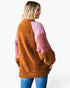 A woman wearing a brown teddy fleece cardigan with pink color-blocked shoulder and sleeve panels, paired with blue jeans, shown from a three-quarters back view against a white studio background.
