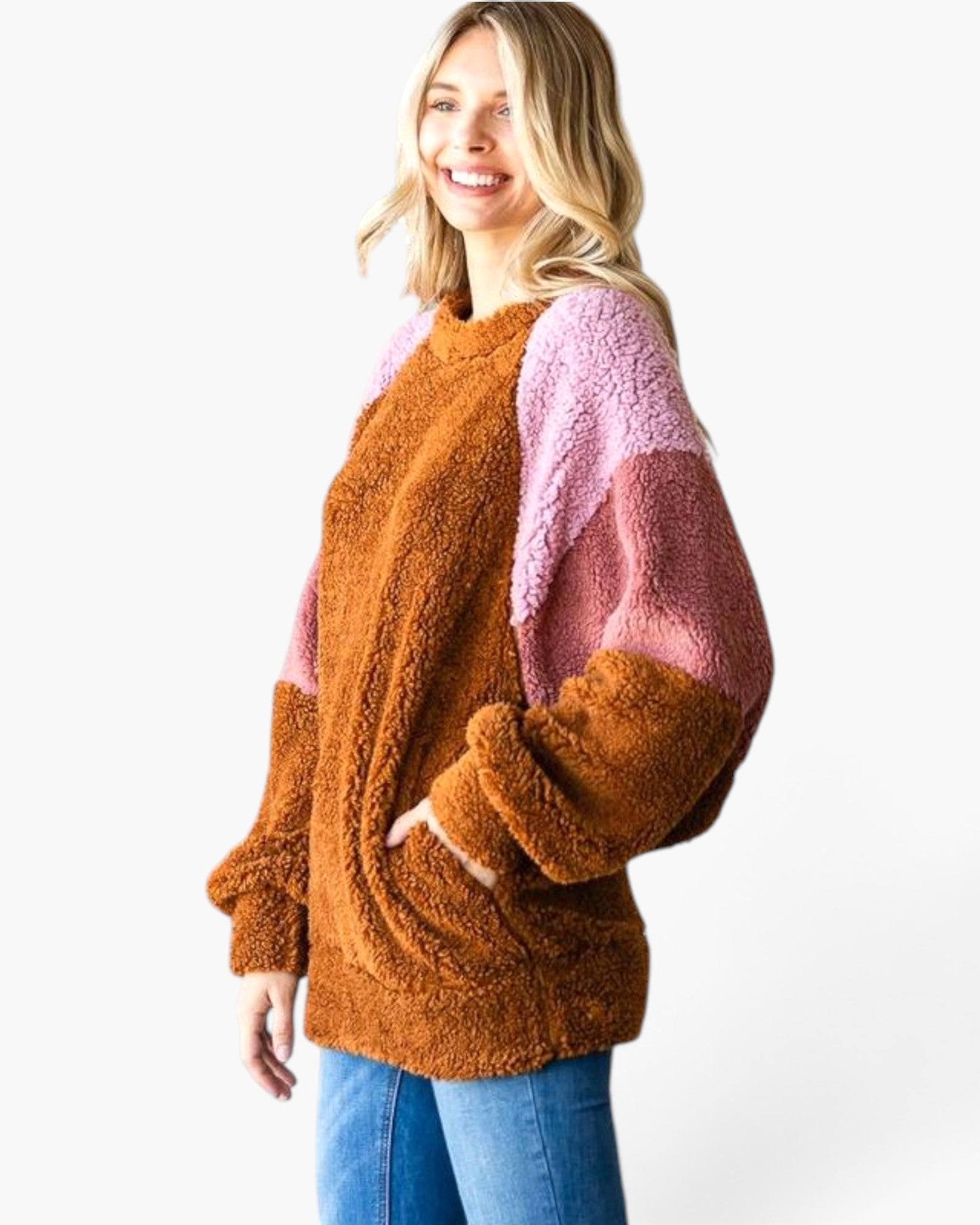 A woman wearing a fuzzy colorblock teddy sweater with a brown body and pink sleeves, paired with blue jeans, against a plain white background.