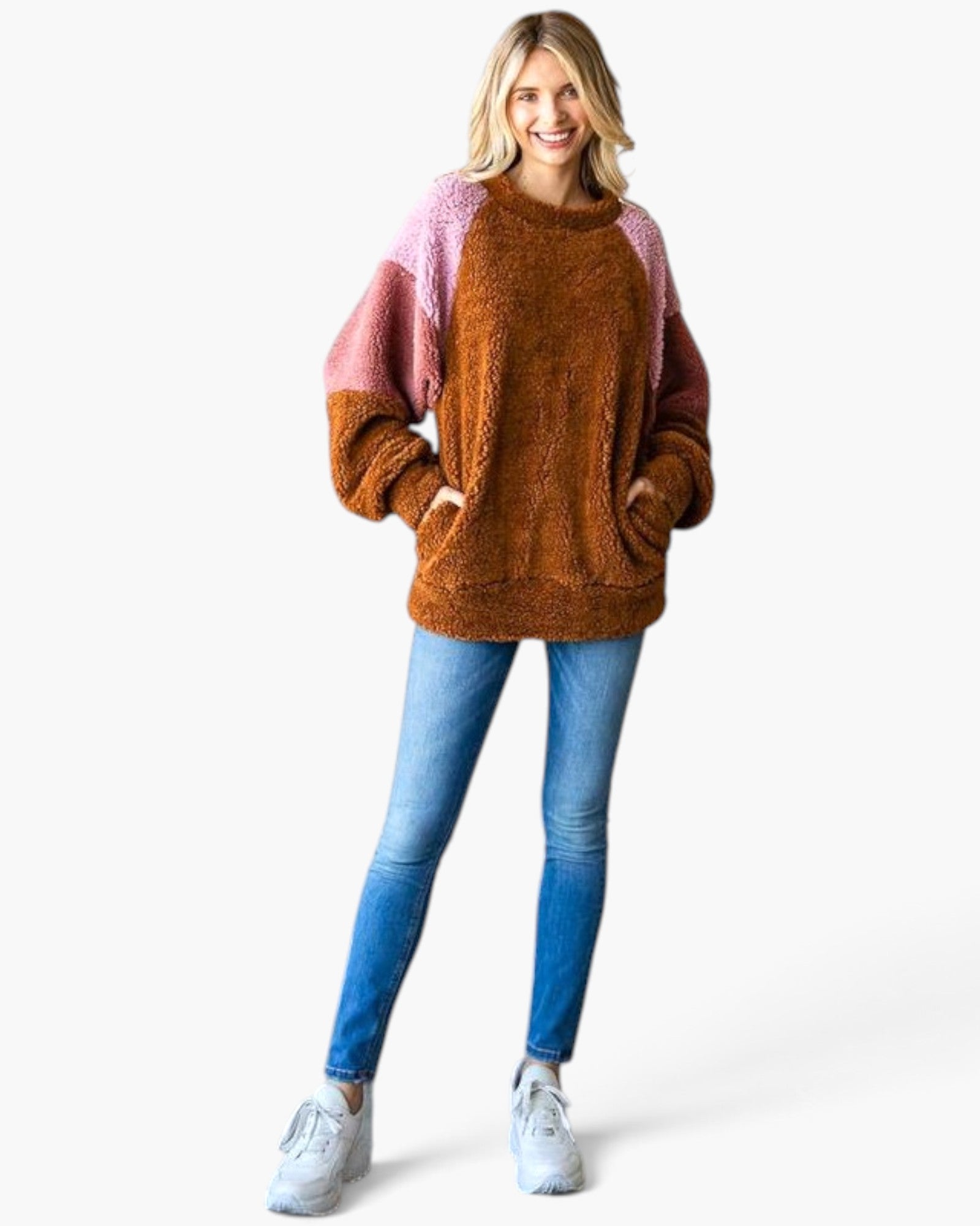 A woman modeling a fuzzy colorblock crewneck sweater with a brown body, pink sleeves, and pink shoulder yokes, paired with blue jeans and white sneakers against a white studio background.