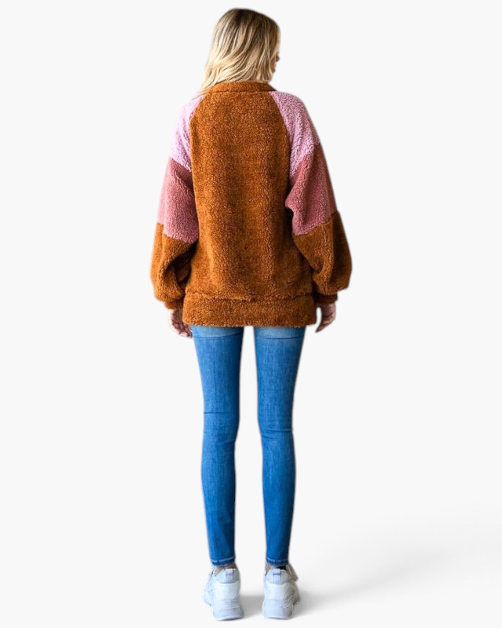 A woman viewed from behind wearing a fuzzy, color-blocked knit sweater with a brown body and pink sleeves and upper back, paired with blue skinny jeans and white sneakers, against a plain white background.