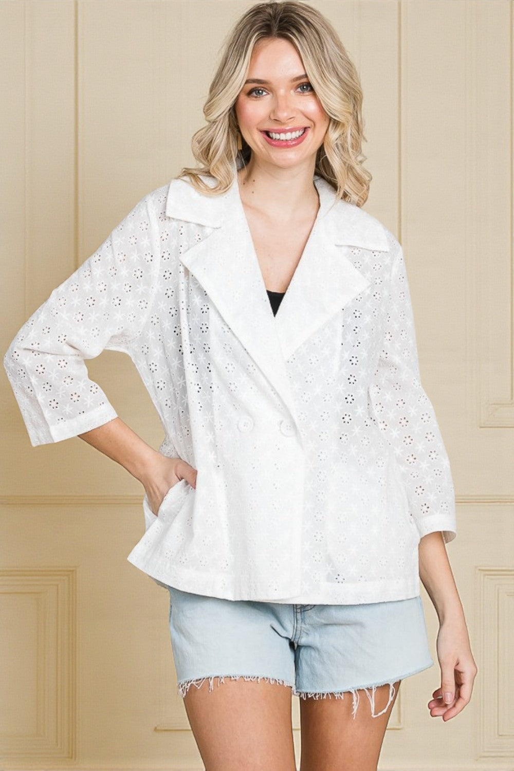 Woman wearing a white embroidered top and light blue shorts against a beige wall.