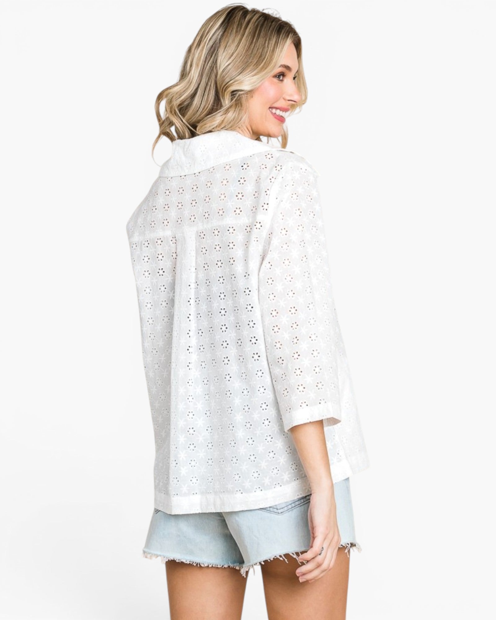 A woman shown from the back wearing a white eyelet button-up shirt and light denim shorts against a plain white studio background.