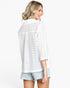 A woman shown from the back wearing a white eyelet button-up shirt and light denim shorts against a plain white studio background.