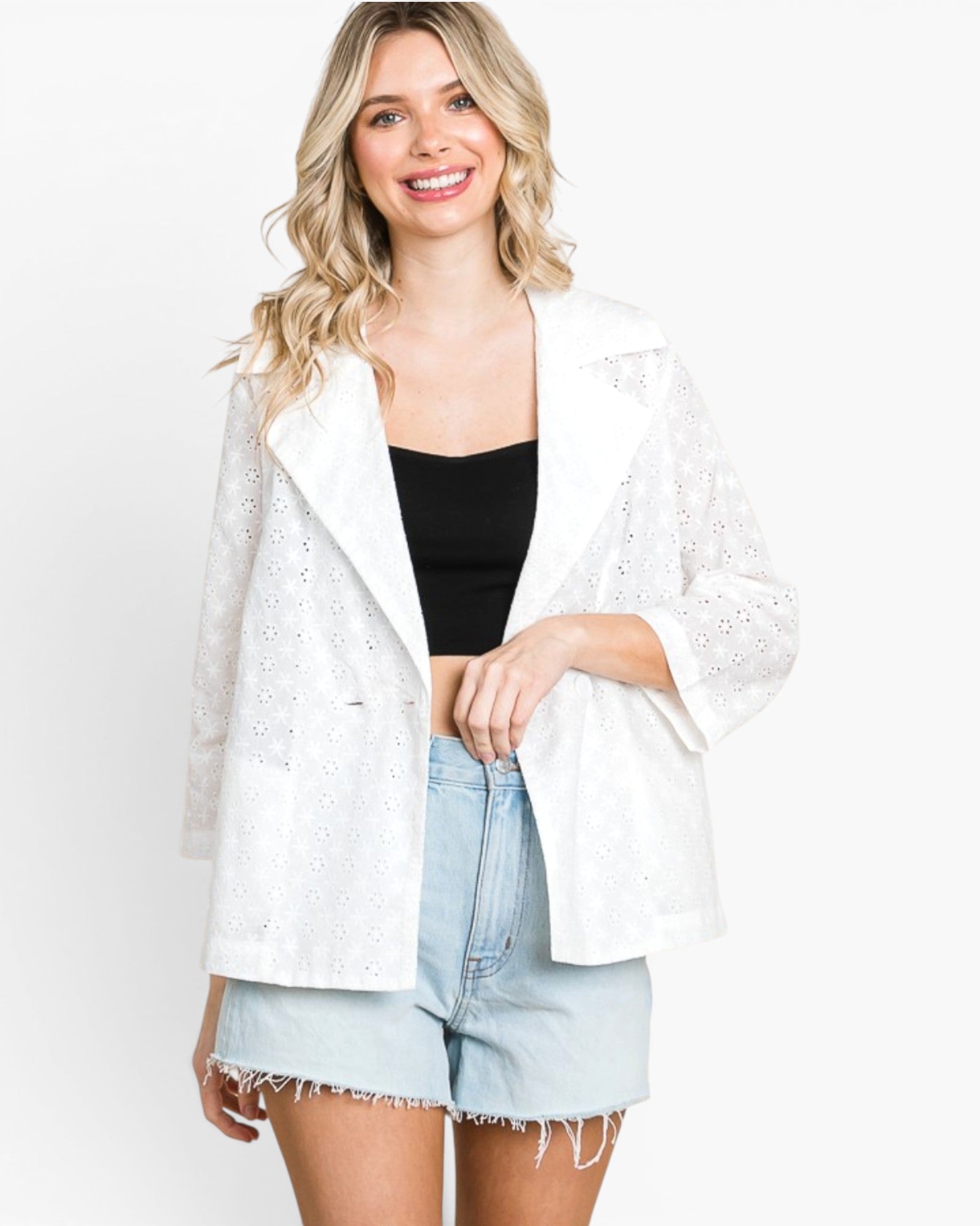 A woman modeling a white eyelet lace open-front jacket over a black tank top and light-wash denim shorts against a plain white studio background.