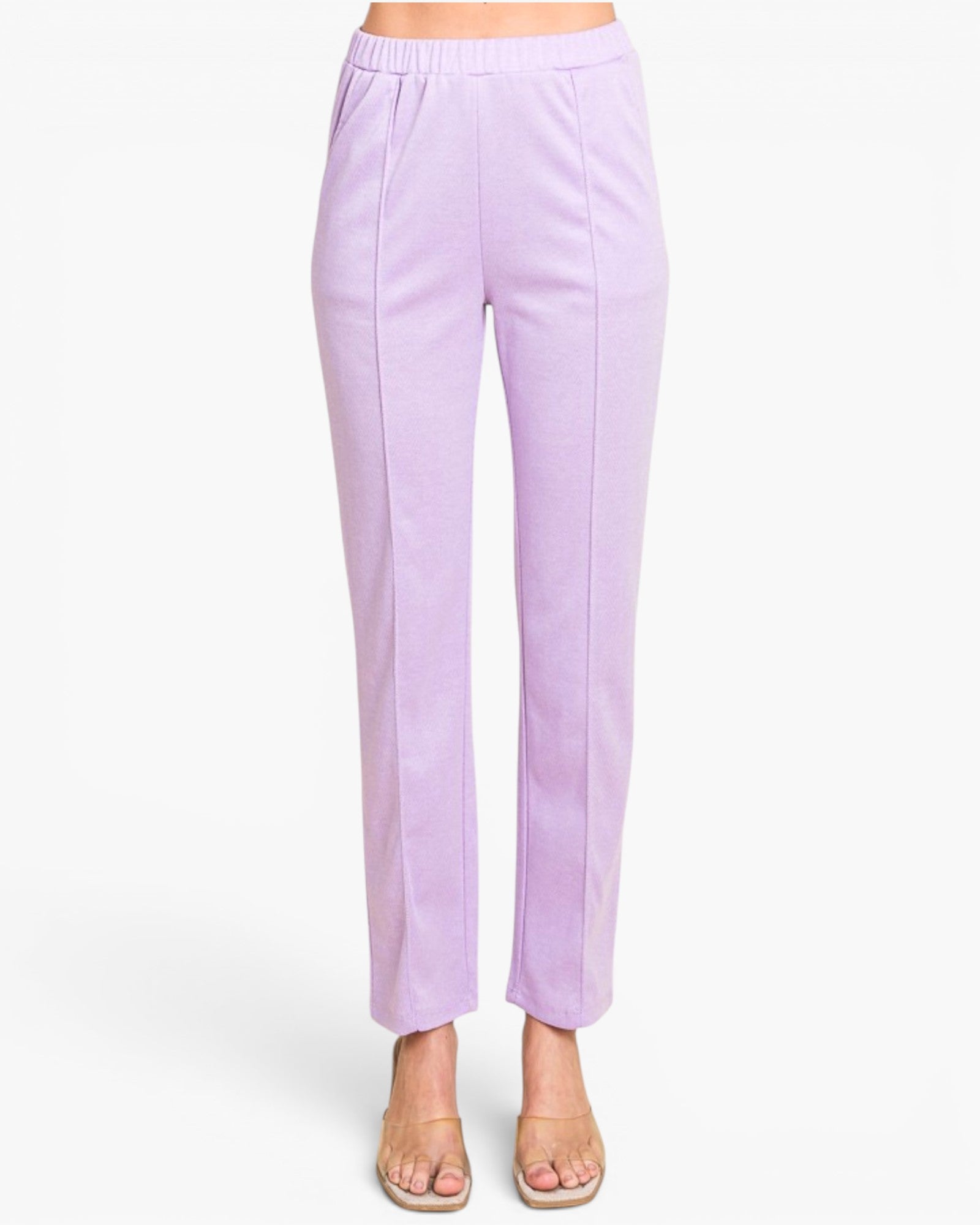 A model is wearing a pair of light purple cropped pants with a wide elastic waistband and side pleats, paired with beige flat sandals against a plain white background.