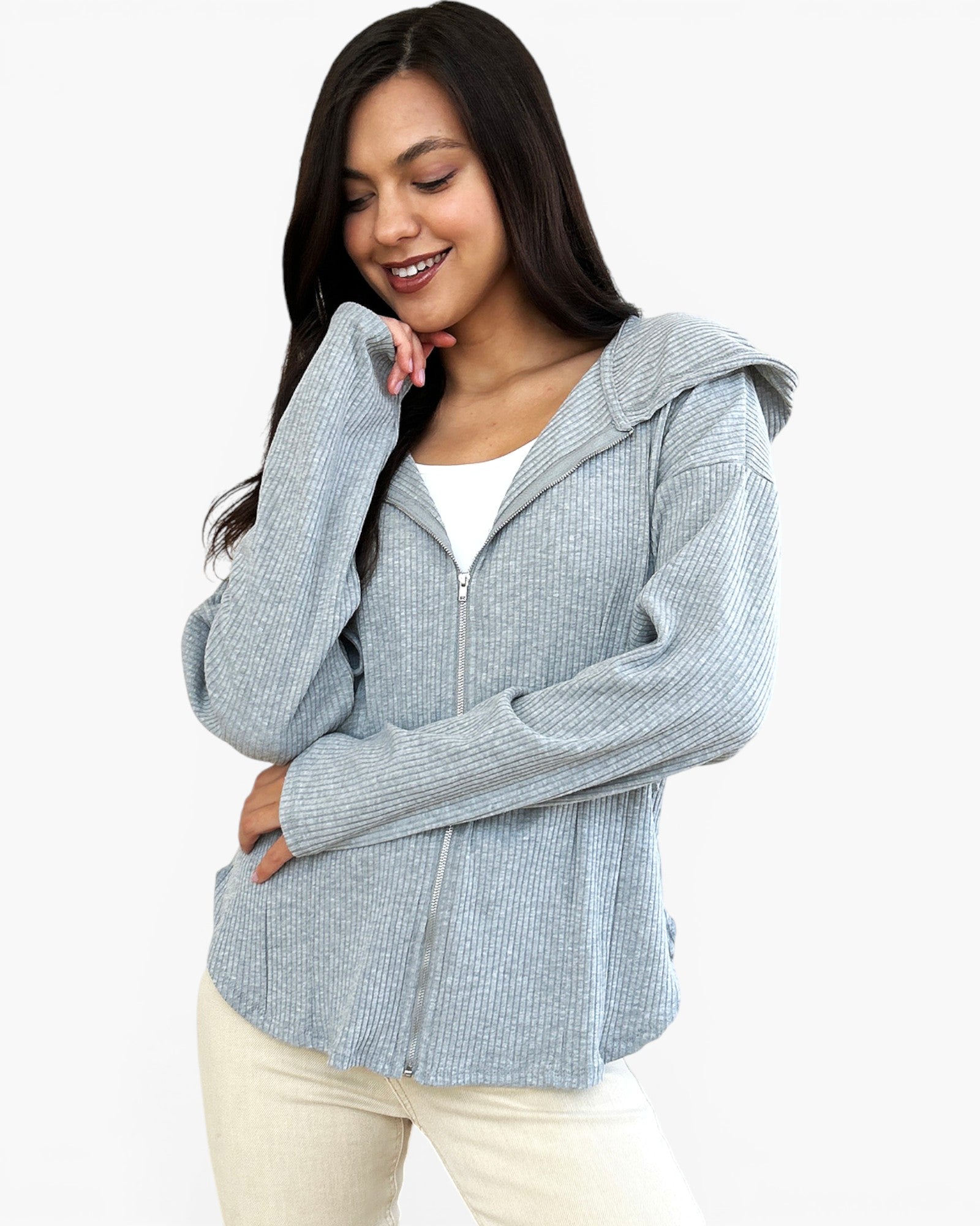 A woman wearing a light blue-gray ribbed zip-up hooded sweater over a white top and cream pants, photographed against a plain light gray studio background.