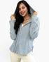 A woman wearing a light blue-gray ribbed zip-up hoodie with the hood up, paired with cream pants, against a plain white background.