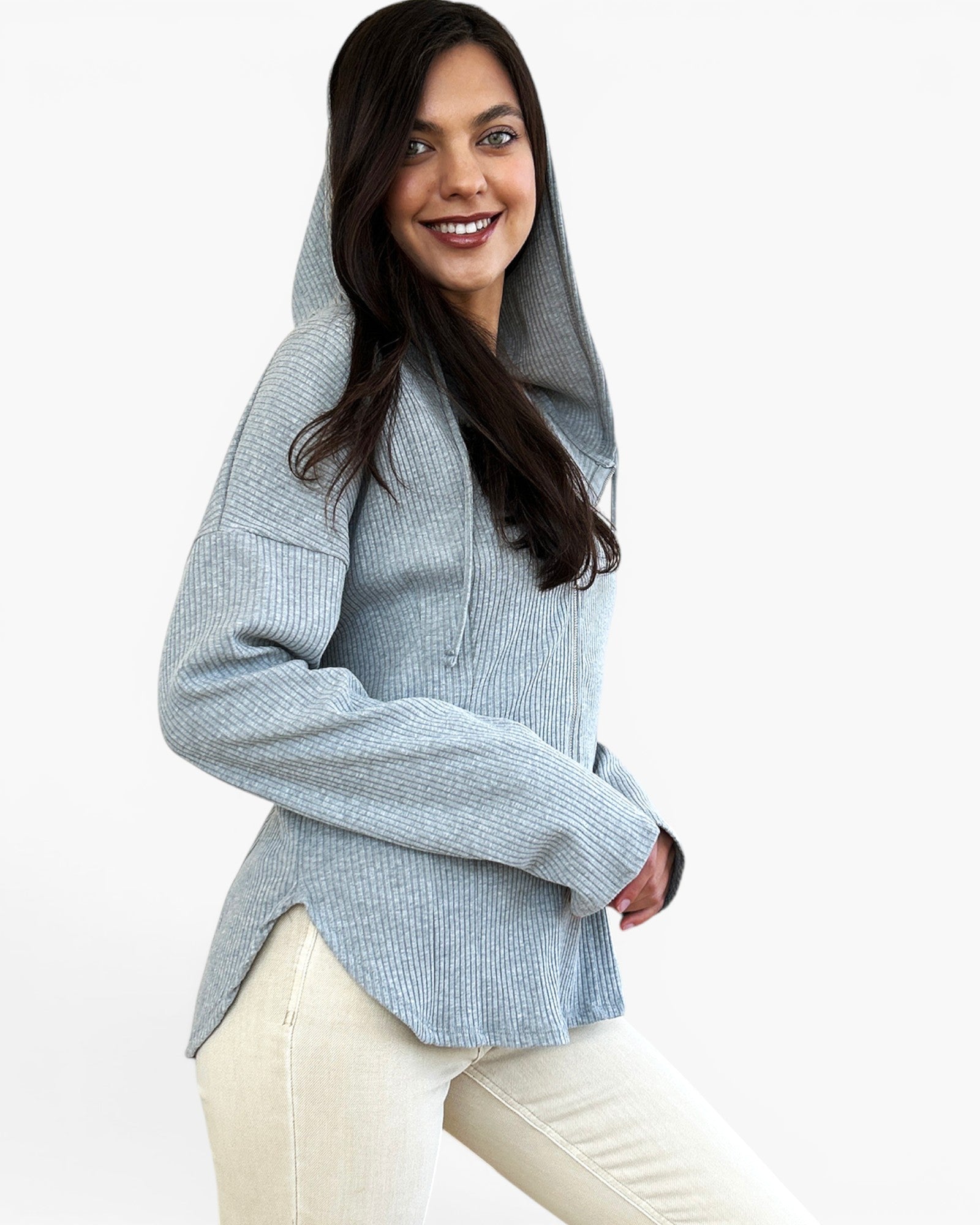 A woman wearing a light blue-gray ribbed knit hooded sweatshirt with a high collar and long sleeves, paired with cream pants, against a plain white studio background.