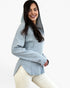 A woman wearing a light blue-gray ribbed knit hooded sweatshirt with a high collar and long sleeves, paired with cream pants, against a plain white studio background.