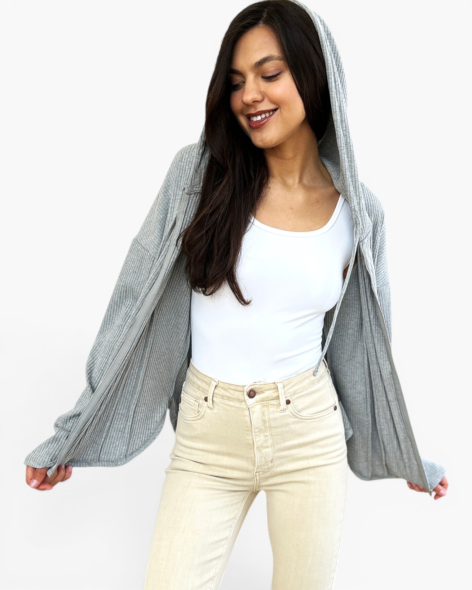 A woman wearing a light gray ribbed knit hooded cardigan over a white tank top and cream-colored jeans, posing against a solid white background.