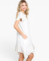 A woman modeling a white short-sleeve dress with ruffle details and side pockets, photographed against a clean white studio background.